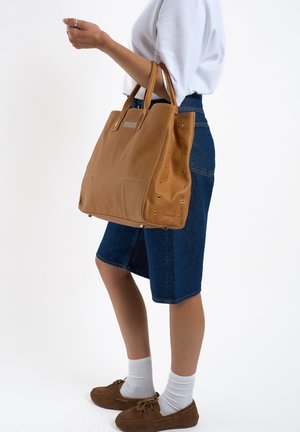 Sac fourre-tout en cuir tan avec deux poignées supérieures, présentant des accents d'étoile sur les côtés et une forme rectangulaire. Jupe en denim et chaussures brunes en arrière-plan.