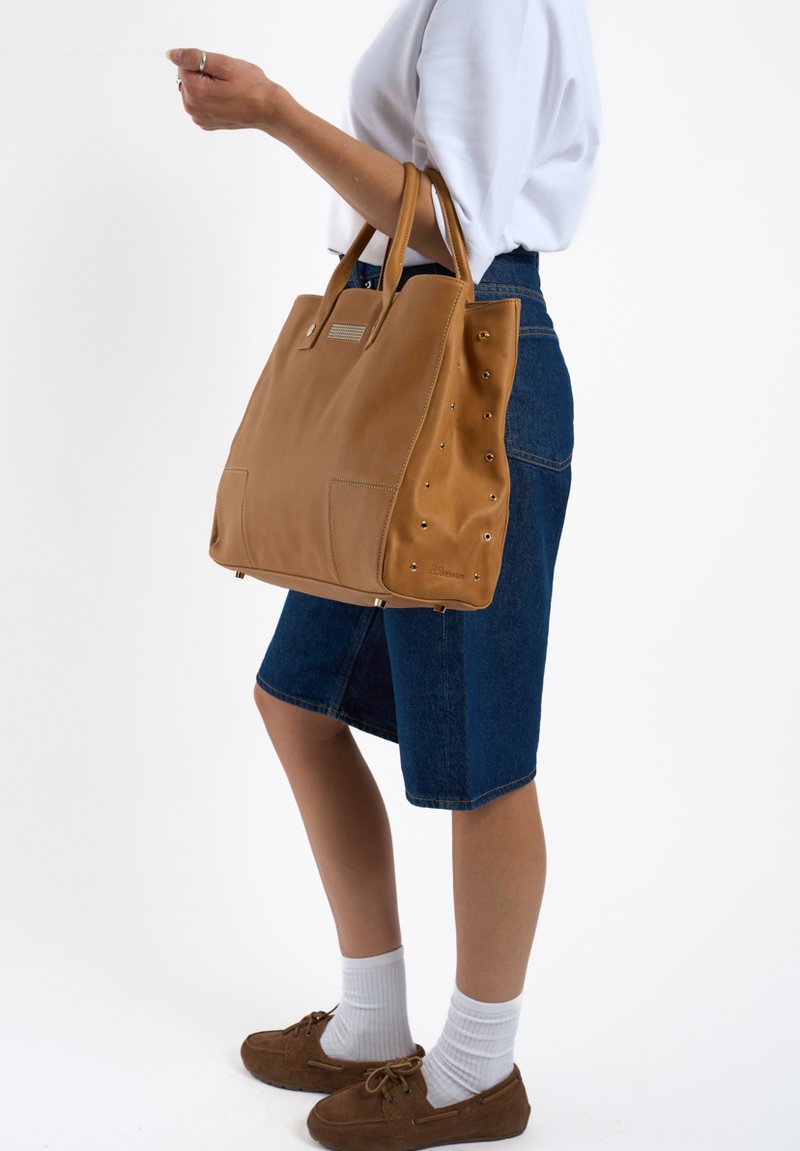 Sac fourre-tout en cuir tan avec deux poignées supérieures, présentant des accents d'étoile sur les côtés et une forme rectangulaire. Jupe en denim et chaussures brunes en arrière-plan.