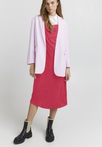 Blazer oversize rosa su un abito a sottoveste rosso, abbinato a stivaletti neri con accenti beige. Tessuto morbido con un design elegante.