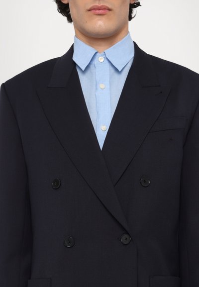 Blazer bleu marine croisé avec des boutons noirs, des revers pointus et une poche poitrine, associé à une chemise bleu clair à boutons.
