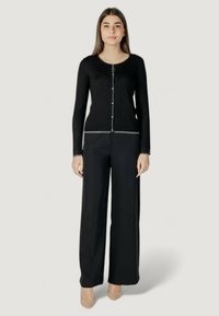Cardigan nero in maglia con scollatura rotonda, rifiniture argentate e bottoni. Abbinato a pantaloni neri a gamba larga e tacchi di colore neutro.
