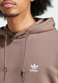 Detailní záběr osoby nosící hnědou mikinu Adidas s bílým logem a šňůrkami, zobrazující část jejich tváře a kudrnaté vlasy.