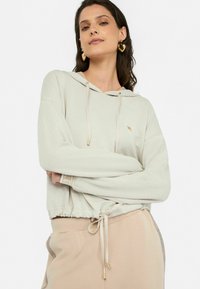 Sudadera recortada de color gris claro con un bolsillo frontal, cuerdas en la capucha y detalles en oro. Combinada con pantalones de chándal beige con cordones.