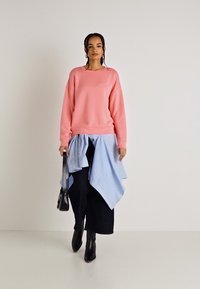 Rosa hettegenser, blåstripet skjorte knyttet rundt livet, svarte vide bukser og svarte hælboots. Glatt stoff og avslappet passform.