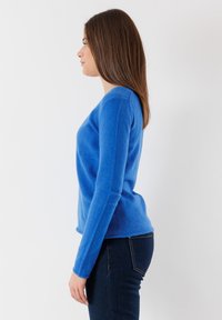 Maglione blu a maniche lunghe in morbido tessuto testurizzato con scollatura rotonda, silhouette aderente e orlo leggermente sfilacciato. Indossato con jeans scuri.