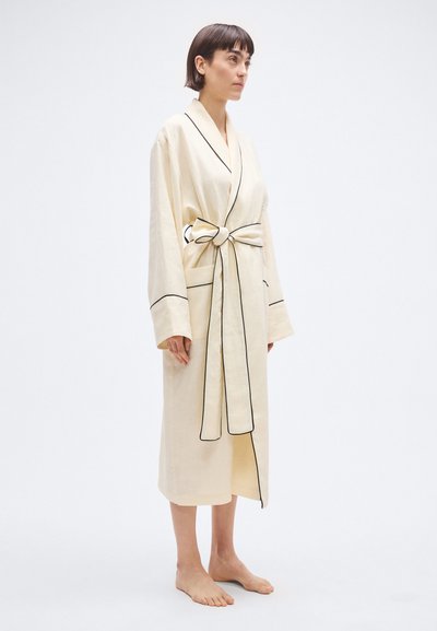 Bēša halāts ar jostu un melnām maliņām, izgatavots no viegla auduma. Raksturo garas piedurknes un ceļgala garuma dizains.