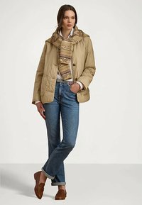 Sieviete, kas ģērbusies bēšā quiltētā jaciņā ar rakstainu šalli, baltā krekla, zilo džinsu un brūnām loafer kedām, stāv ar vienu roku kabatā.