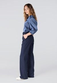 Blauwe satijnen blouse met pofmouwen, gecombineerd met marineblauwe pantalon met streepjes en brede pijpen. Model draagt witte sneakers en heeft haar handen in de zakken.