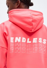 Persoană cu părul împletit purtând un hanorac roz aprins cu cuvântul „ENDLESS” imprimat în alb, repetat în text stratificat pe spate.
