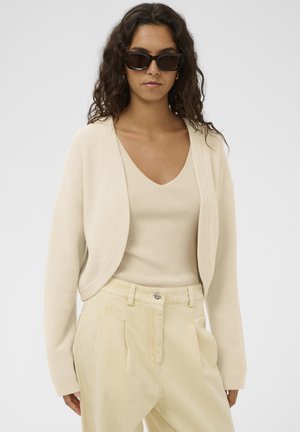 Femme portant des lunettes de soleil foncées, un cardigan court crème, un haut crème à col en V et un pantalon taille haute beige clair sur un fond uni.