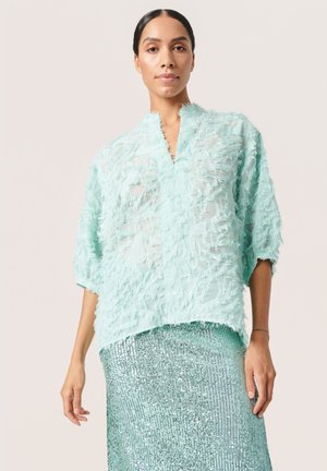 Blusa de un aqua claro con un tejido texturizado y deshilachado, y un diseño de cuello en V, combinada con una falda de lentejuelas plateadas brillantes. Mangas cortas y corte holgado.