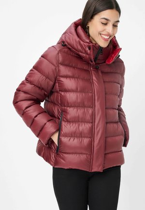Frau lächelt, trägt eine bordeauxrote gesteppten Steppjacke mit hohem Kragen, Kapuze und seitlichen Reißverschlusstaschen, die Hände in den Taschen.