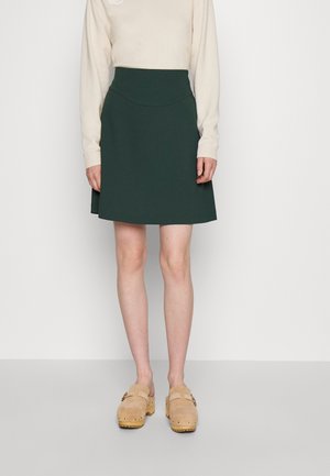 Vrouw draagt een donkergroene knie-lange rok, een beige top met lange mouwen en beige suède klompen met houten zolen, staand tegen een effen achtergrond.