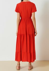 Robe maxi rouge avec des manches courtes en volants, taille élastique, jupe à volants et détail en boutons au dos. Portée avec des sandales métalliques.