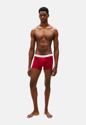 Modelo masculino de pie descalzo, vistiendo bóxers rojos de Tommy Hilfiger con una cinturilla blanca con la marca, sobre un fondo liso.