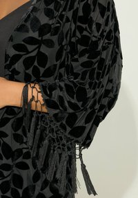 Svart kimono-liknande jacka med texturerat lövmönster, sammetstyg, lös passform och fransdetaljer längs ärmarna för extra detalj.