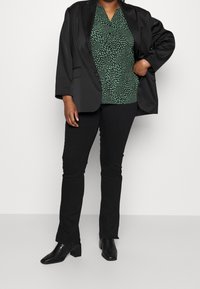 Blazer nero sopra una blusa a fantasia verde, abbinato a pantaloni neri aderenti e scarpe con tacco nere. Tessuto morbido, vestibilità rilassata e scollatura a V.
