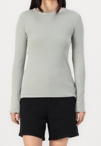 Osoba v světle šedém dlouhém rukávu s žebrováním a černých šortkách stojící na pozadí s bílou plochou.