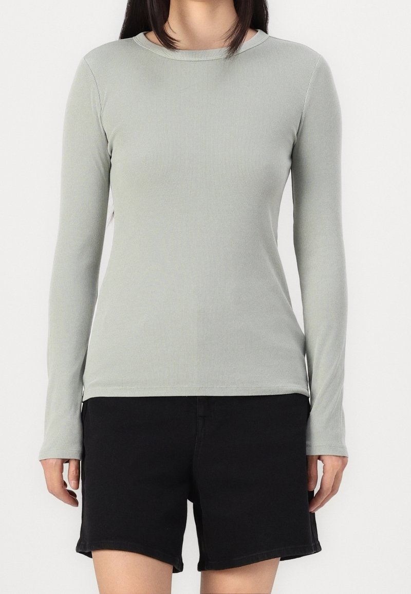 Osoba v světle šedém dlouhém rukávu s žebrováním a černých šortkách stojící na pozadí s bílou plochou.