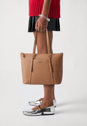 Borsa tote color beige con due manici, superficie testurizzata e stampa del logo. Presenta una forma rettangolare e chiusura con zip. Indossata con scarpe basse argentate.