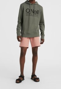 Felpa verde oliva con logo nero, pantaloni corti rosa e sandali neri. La felpa presenta un cappuccio con coulisse e polsini a costine. I pantaloni corti sono di lunghezza al ginocchio.