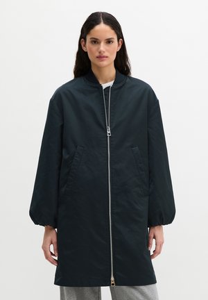 Persoon met lang donker haar, gekleed in een lang zwart jack met ritssluiting, elastische manchetten en schuine zakken aan de voorkant, staand tegen een effen achtergrond.