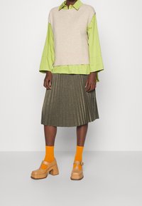 Gilet lavorato a maglia beige sopra una camicia verde brillante con colletto, gonna plissettata oliva e scarpe con plateau color sabbia abbinate a calzini arancioni.