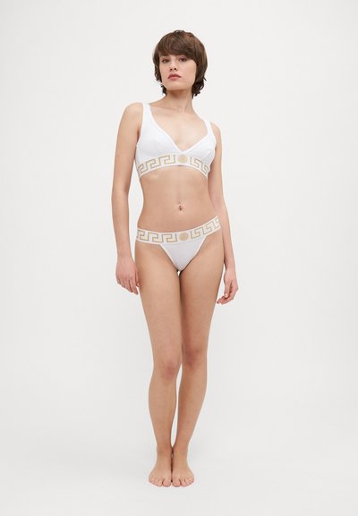 Balta bikini komplekts, kas ietver v-veida krūšturi ar zelta ģeometrisku rakstu ap joslu un saskaņoto stringu. Gluda, stieptspējīga auduma.