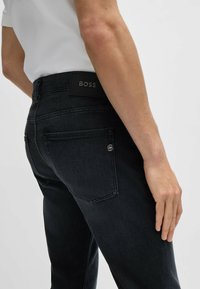 Dark denim jeans with a fitted design, featuring a black leather logo patch at the waistband, subtle stitching, and two back pockets.