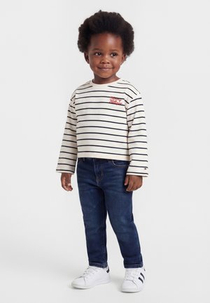 Małe dziecko z kręconymi włosami, ubrane w białą, długorękawową koszulkę w paski, ciemnoniebieskie dżinsy i białe trampki, stoi i się uśmiecha.