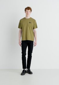 Camiseta verde oliva con "LEVI'S" impreso en negro, combinada con jeans ajustados negros y zapatos negros de cordones, sobre un fondo neutro.