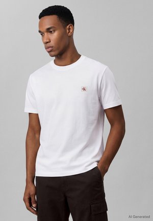 Fiatal férfi rövid, göndör hajjal, egyszerű fehér pólót visel kis mellkas logóval és sötét cargo nadrággal, szürke háttér előtt áll.