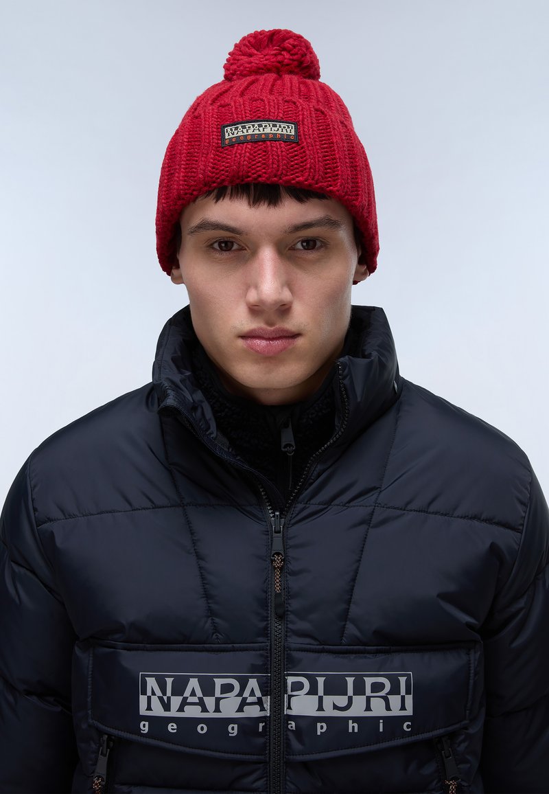 Beanie rosso lavorato a maglia con pom-pom superiore e una toppa nera con logo, indossato con una giacca imbottita nera con zip che presenta un logo sul petto.