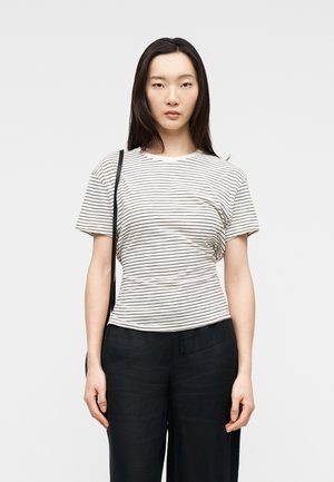 Ung kvinde med glat mørkt hår iført en hvid kortærmet skjorte med striber, sorte bukser og en sort skuldertaske, stående mod en ensfarvet baggrund.