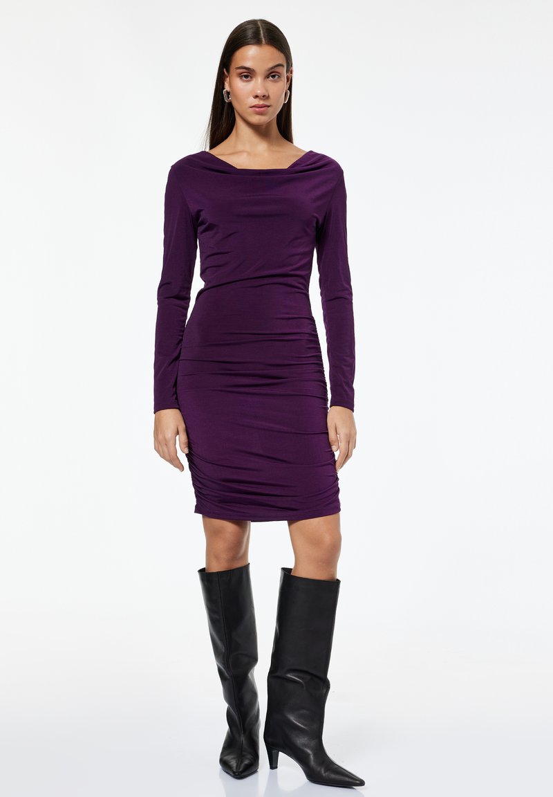 Donna in piedi che indossa un abito lungo a maniche lunghe di colore viola con arricciature e stivali con tacco neri fino al polpaccio su uno sfondo bianco.