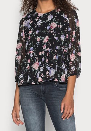 Vrouw die een zwarte doorschijnende blouse met bloemenprint draagt, met roze en paarse bloemen, driekwartmouwen, gecombineerd met donkerblauwe jeans.