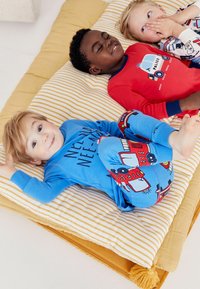 Trois enfants sont allongés sur un matelas rayé. L'un porte une chemise bleue avec du texte et un pantalon bleu à motifs de camions de pompiers, un autre a un haut rouge, et le dernier porte un ensemble de pyjama à motifs.