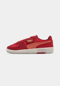 Baskets rouges avec une tige en daim et textile, présentant une semelle en caoutchouc beige, des accents dorés et un logo PUMA texturé sur le côté.