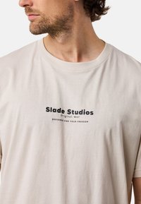 Beige bomulds T-shirt med rund halsudskæring, korte ærmer og trykt tekst i sort: "Slade Studios Original Wear" og "DESIGNED FOR YOUR FREEDOM."