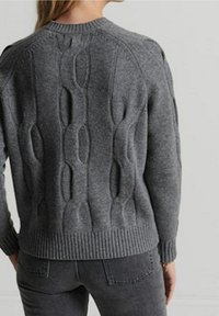 Personne portant un pull en maille gris foncé avec un motif torsadé texturé dans le dos et un jean en denim gris foncé.