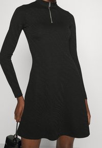 Robe noire à manches longues avec un col montant et une fermeture éclair ; présente un motif texturé subtil sur tout le tissu et une silhouette évasée.
