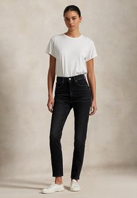 Frau trägt ein weißes T-Shirt mit kurzen Ärmeln, hoch taillierte schwarze Jeans und weiße Turnschuhe und steht vor einem neutralen Hintergrund.
