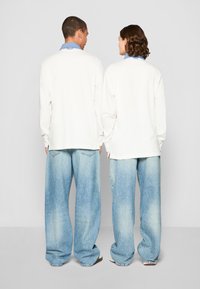 Camisa unisex de manga larga en color crema con un cuello azul, combinada con unos vaqueros sueltos de color azul claro. Tejido suave y corte relajado.