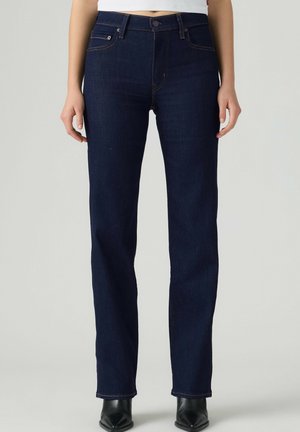 Person trägt dunkelblaue Jeans mit geradem Bein, weißes kurzes Oberteil und schwarze spitze Stiefel, steht vor einem schlichten hellen Hintergrund.