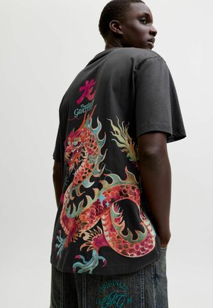 Mladá osoba nosiaca čierne tričko s veľkým farebným motívom draka a textom, doplnené tmavými džínsami.