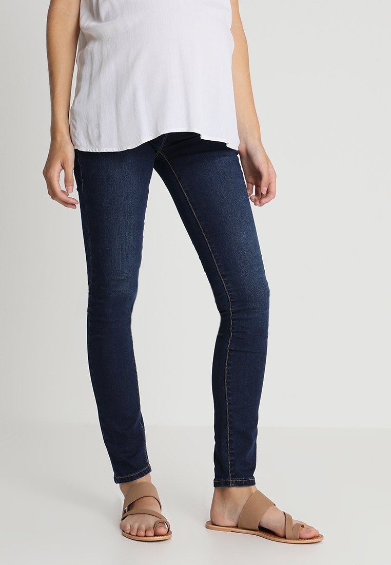 Persona vestida con jeans ajustados azul oscuro, una blusa blanca y sandalias planas color crema, de pie contra un fondo gris claro.