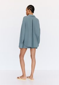 Himmelblaues, strukturiertes Hemd mit langen Ärmeln, seitlichen Schlitzern und Knopfmanschetten, kombiniert mit passenden Shorts und hellen Sandalen. Schlichtes, lässiges Design.