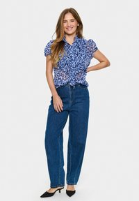 Blusa blu con stampa leopardata e colletto arricciato, abbinata a jeans a vita alta blu scuro e décolleté neri a punta. Tessuto liscio, vestibilità casual.