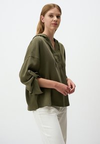 Blusa capuccio verde oliva con vestibilità rilassata, maniche lunghe con dettagli a bottone e scollatura a V. Indossata con pantaloni chiari.