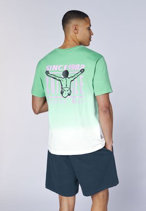 Mann trägt ein T-Shirt mit grün-weißem Farbverlauf, mit einer Grafik von einer Person, die Gewichte hebt, und dem Schriftzug "Since 1982" sowie dunkle Shorts.